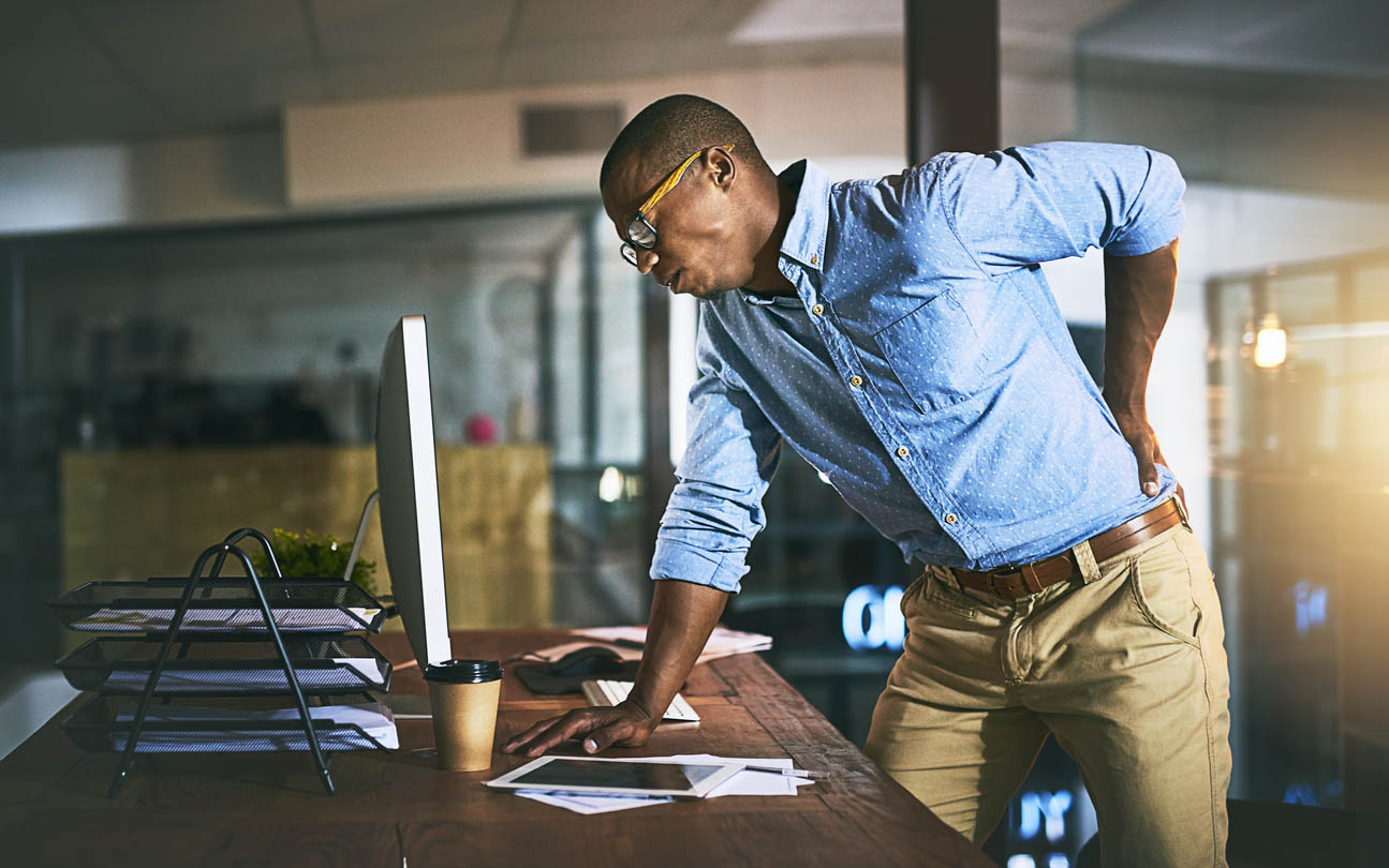 A man standing in front of computer holding his back in pain.
